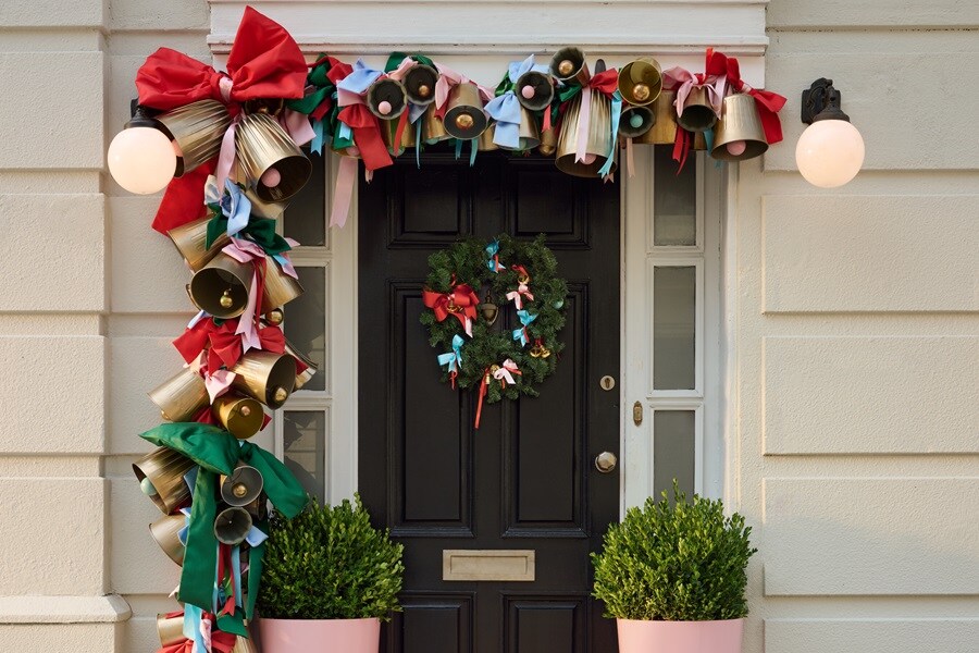 Drzwi wejściowe z girlandą złotych dzwonków i czerwono‑zielonych wstążek zawieszoną na zewnętrznych haczykach Command, a także z wieńcem na haczyku Command do wieńców.
